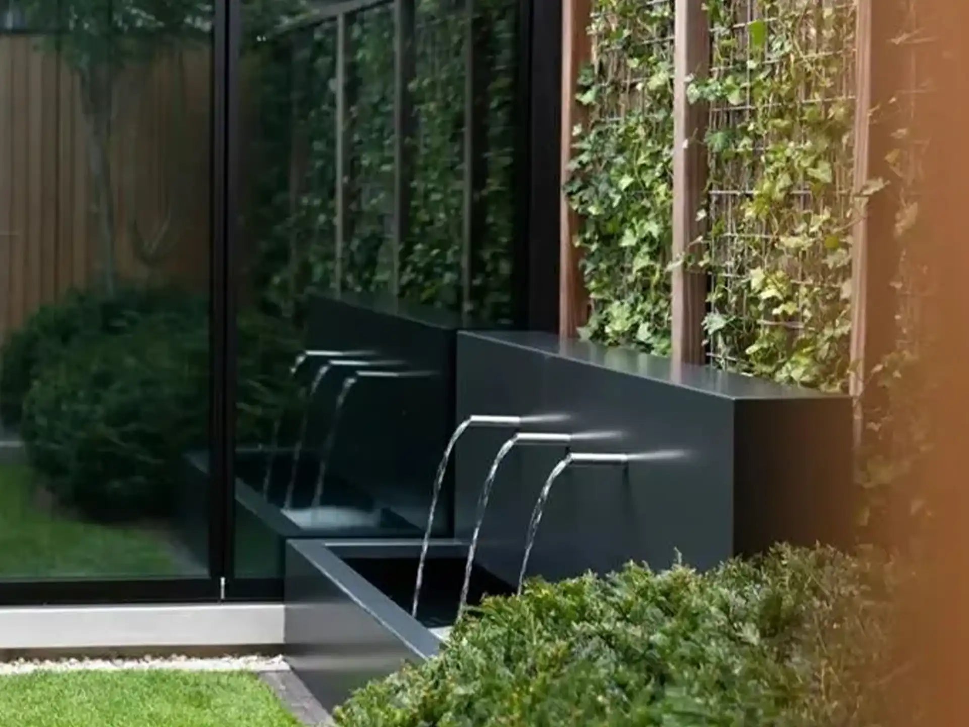 Sfeervolle tuinopstelling met een vrijstaande Adezz vijvermuur van aluminium, voorzien van 3 pijpjes en pomp, geplaatst langs een moderne gevel met glas en een houten schutting, waarbij waterstromen elegant in de vijver vallen.
