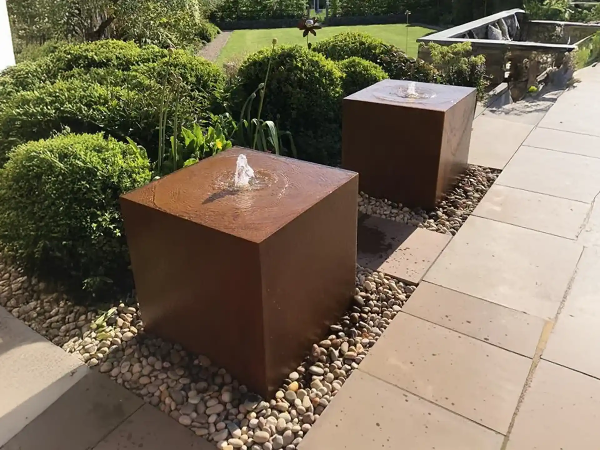 Adezz waterblok van cortenstaal met 3 fonteinen, sfeervol geplaatst in moderne tuin tussen groen en bestrating.