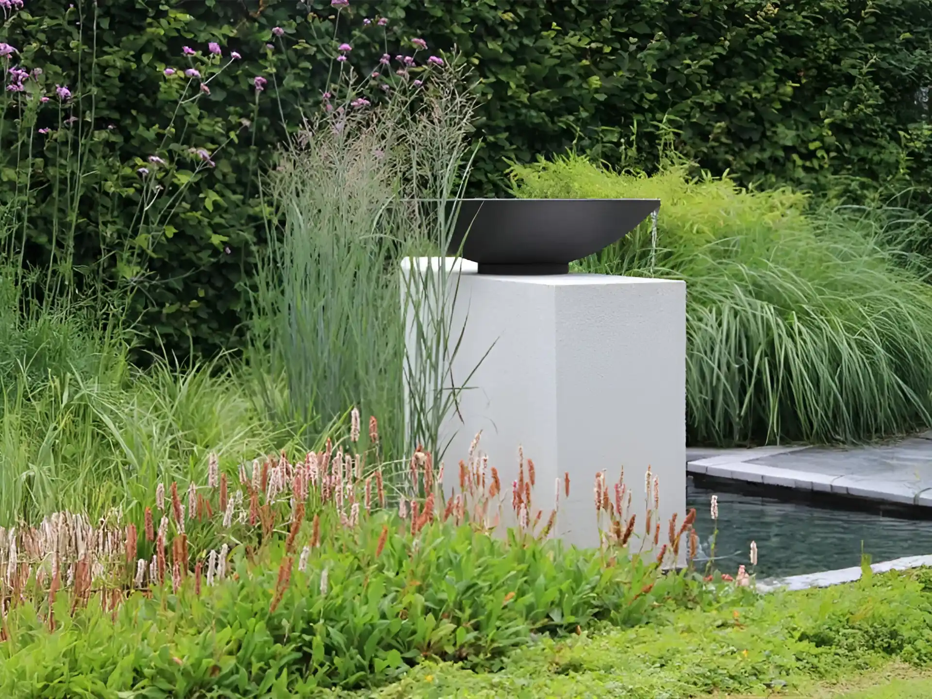 Adezz waterschaal van gecoat staal geplaatst op een witte sokkel, omringd door siergras en planten naast een vijver in de tuin.