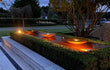 Avondfoto van een cortenstalen Adezz watertafel met vier ronde schalen en geïntegreerde verlichting, die warm oplicht in een stijlvolle tuin met hagen en sierbomen.