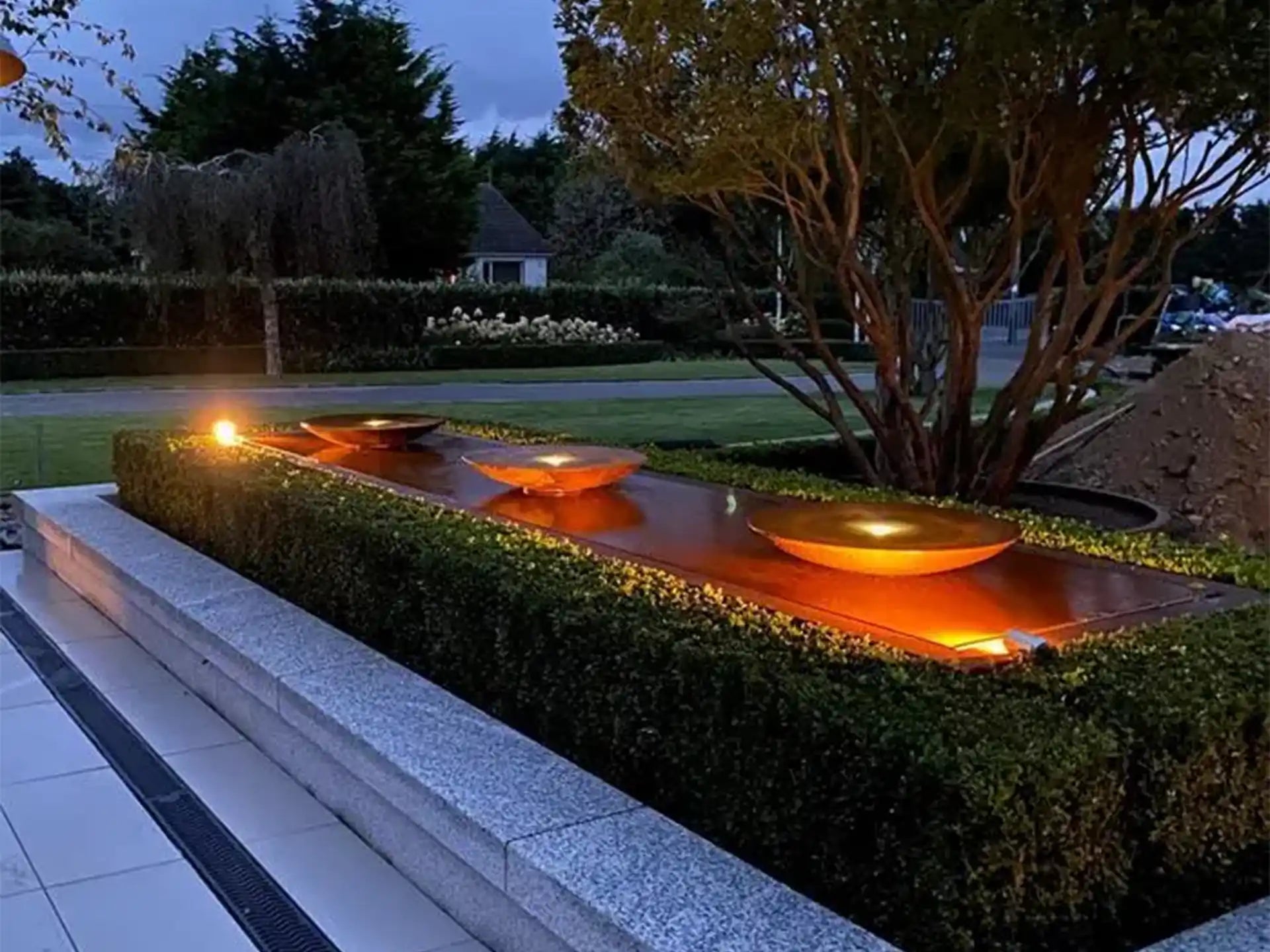 Avondfoto van een cortenstalen Adezz watertafel met vier ronde schalen en geïntegreerde verlichting, die warm oplicht in een stijlvolle tuin met hagen en sierbomen.