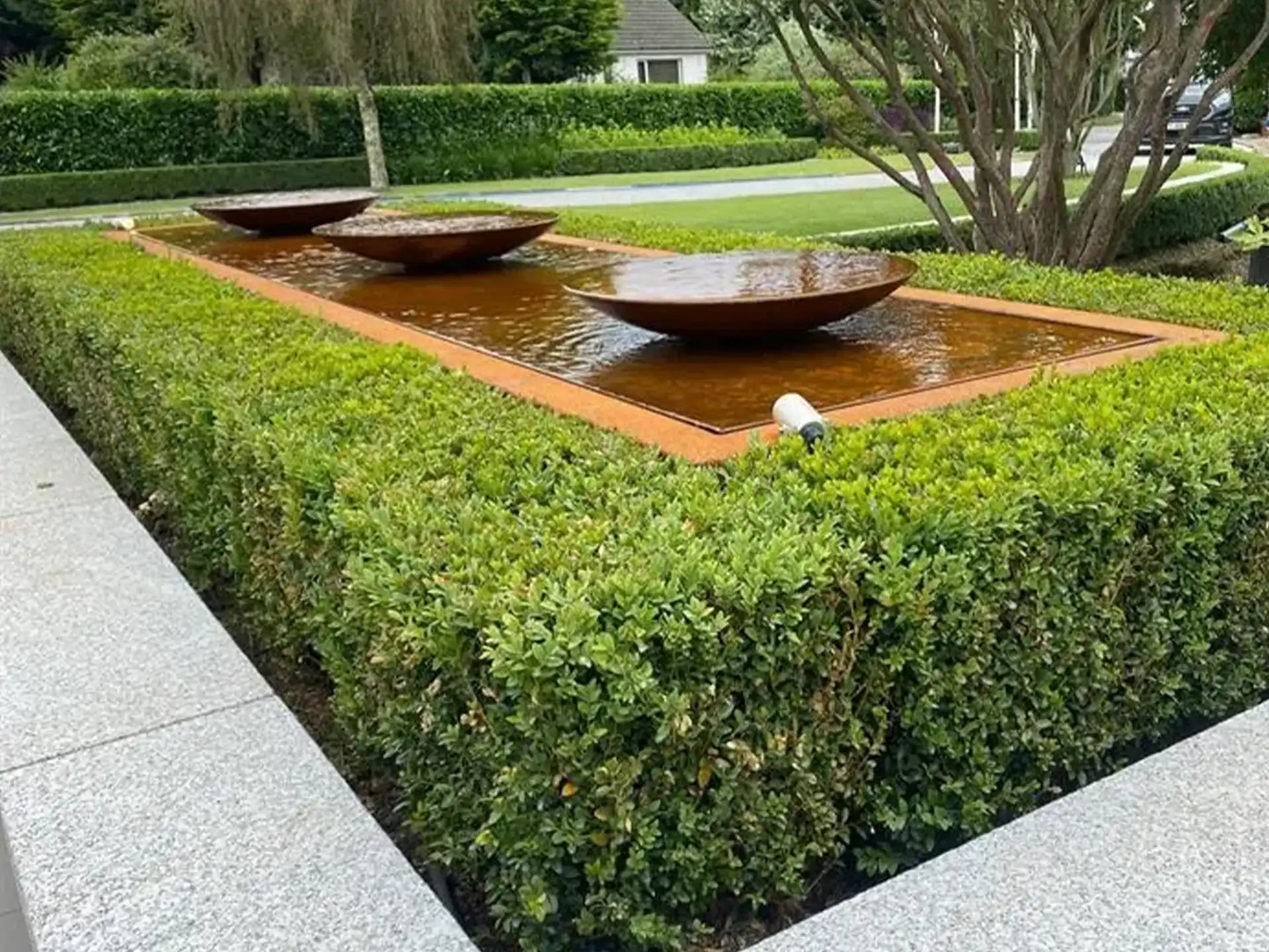 Overdag sfeerbeeld van een cortenstalen Adezz watertafel met drie ronde schalen waar water overheen stroomt, geplaatst in een strakke tuin met lage hagen en moderne bestrating.