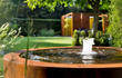 Close-up van Adezz ronde cortenstaal watertafel met fontein, water spuit elegant omhoog in een groene tuin.