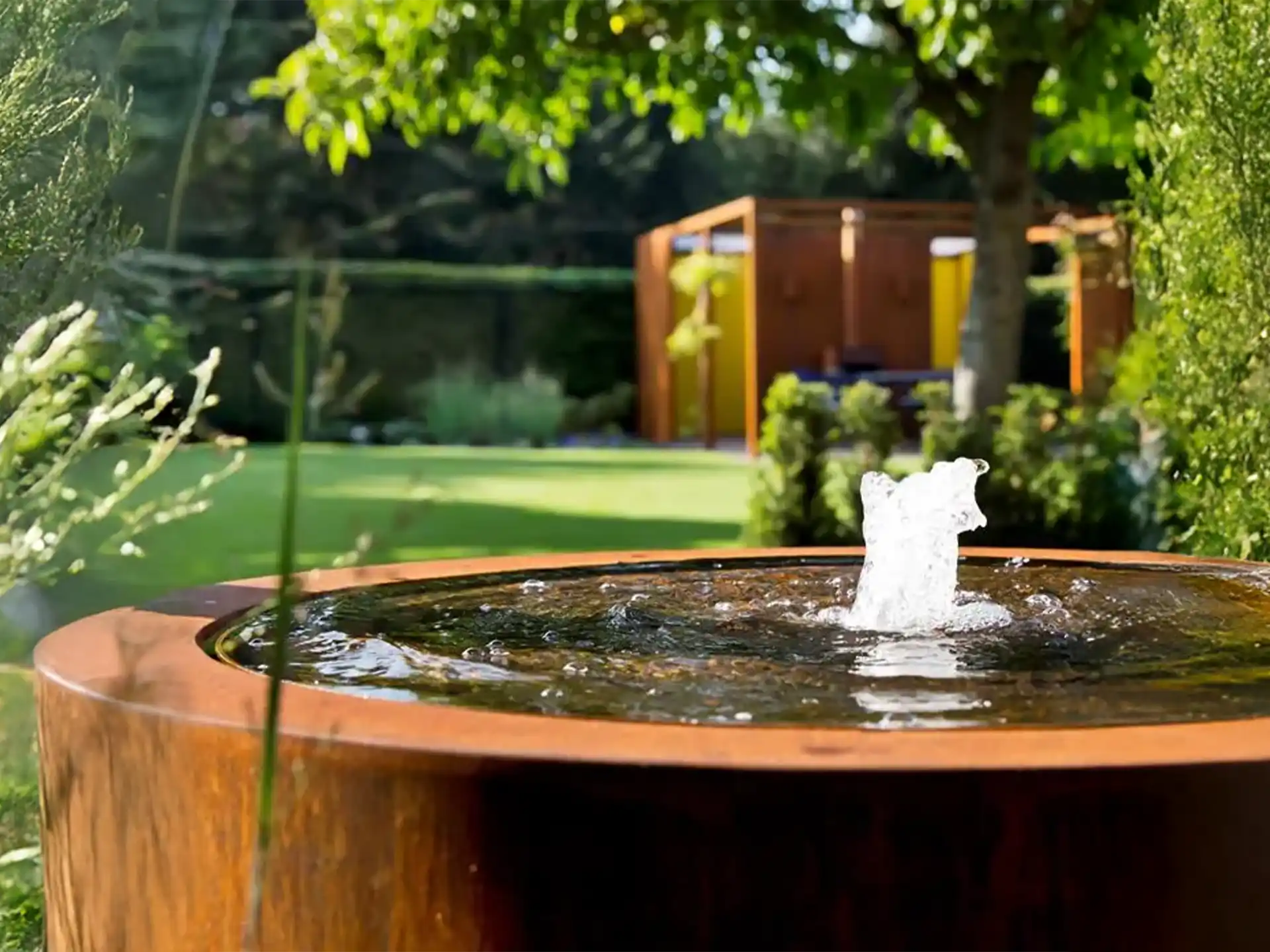 Close-up van Adezz ronde cortenstaal watertafel met fontein, water spuit elegant omhoog in een groene tuin.