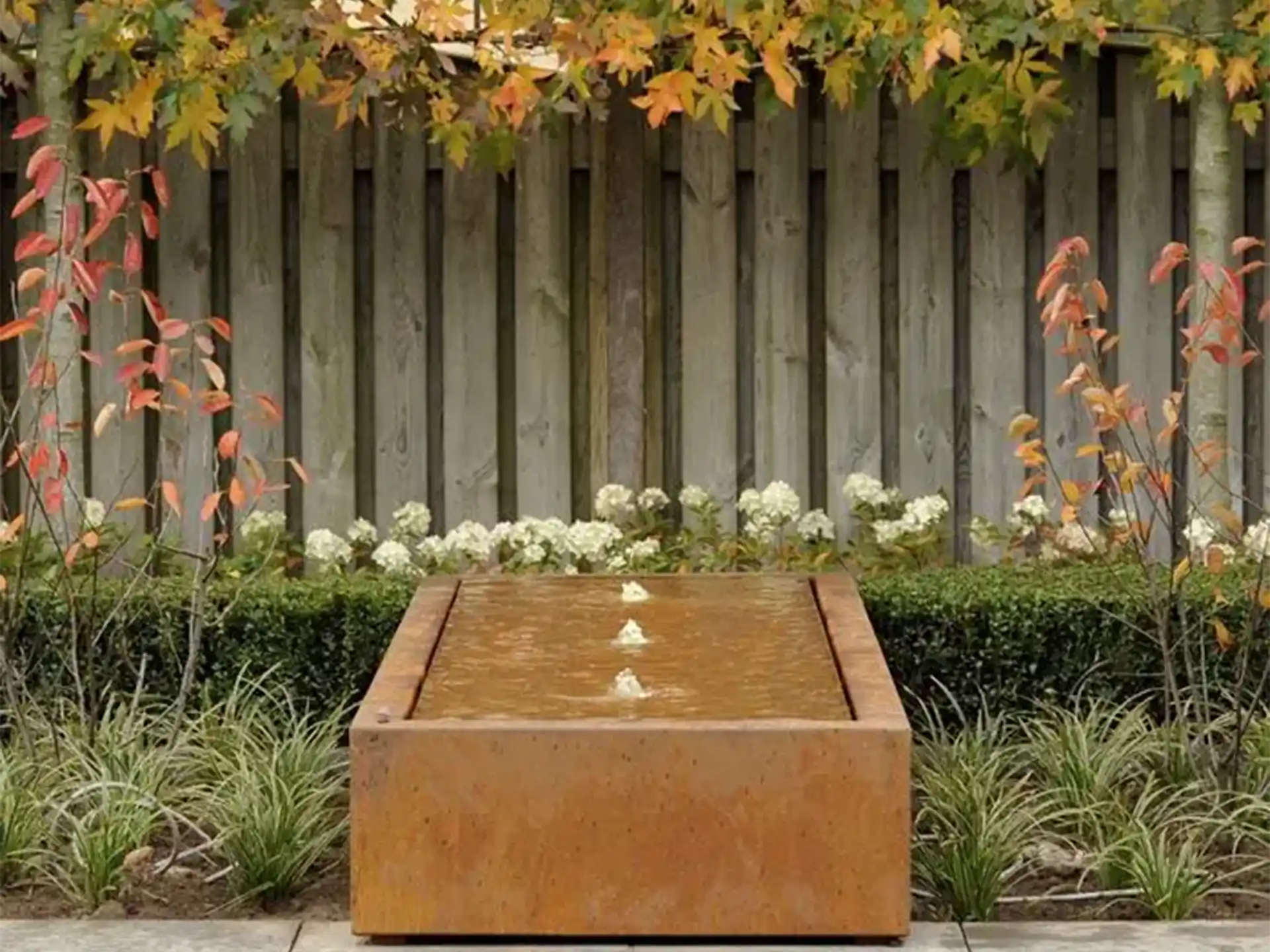 Adezz watertafel van cortenstaal in vierkant model met fontein, geplaatst in een tuin met groene beplanting.