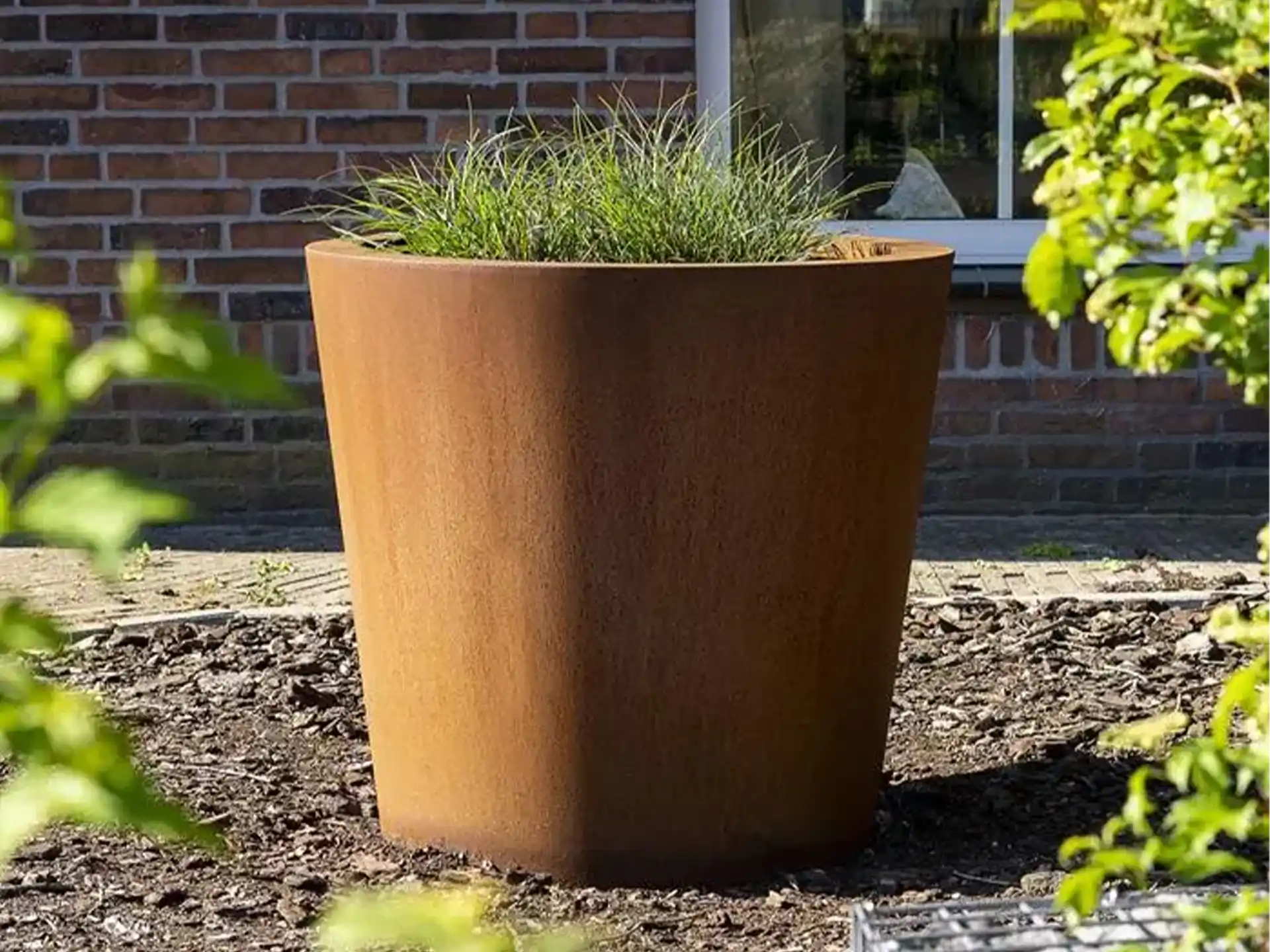 Adezz Conic Cortenstaal Plantenbak Rond met bodem, gevuld met grasplanten naast een woning.