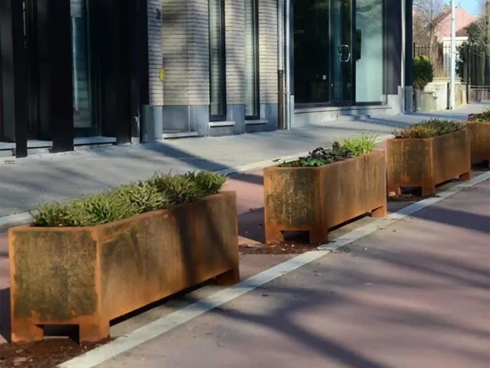 Rij met Adezz Carrez Cortenstaal Plantenbakken Rechthoek met poten en bodem bij een moderne kantoorlocatie.