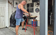 Man in korte broek en hemd bereidt paella in een grote zwarte pan op de rode Garcima paella brander met standaard. De set staat buiten onder een overkapping naast een witte houten wand, met decoratie aan de muur en een gasfles ernaast.