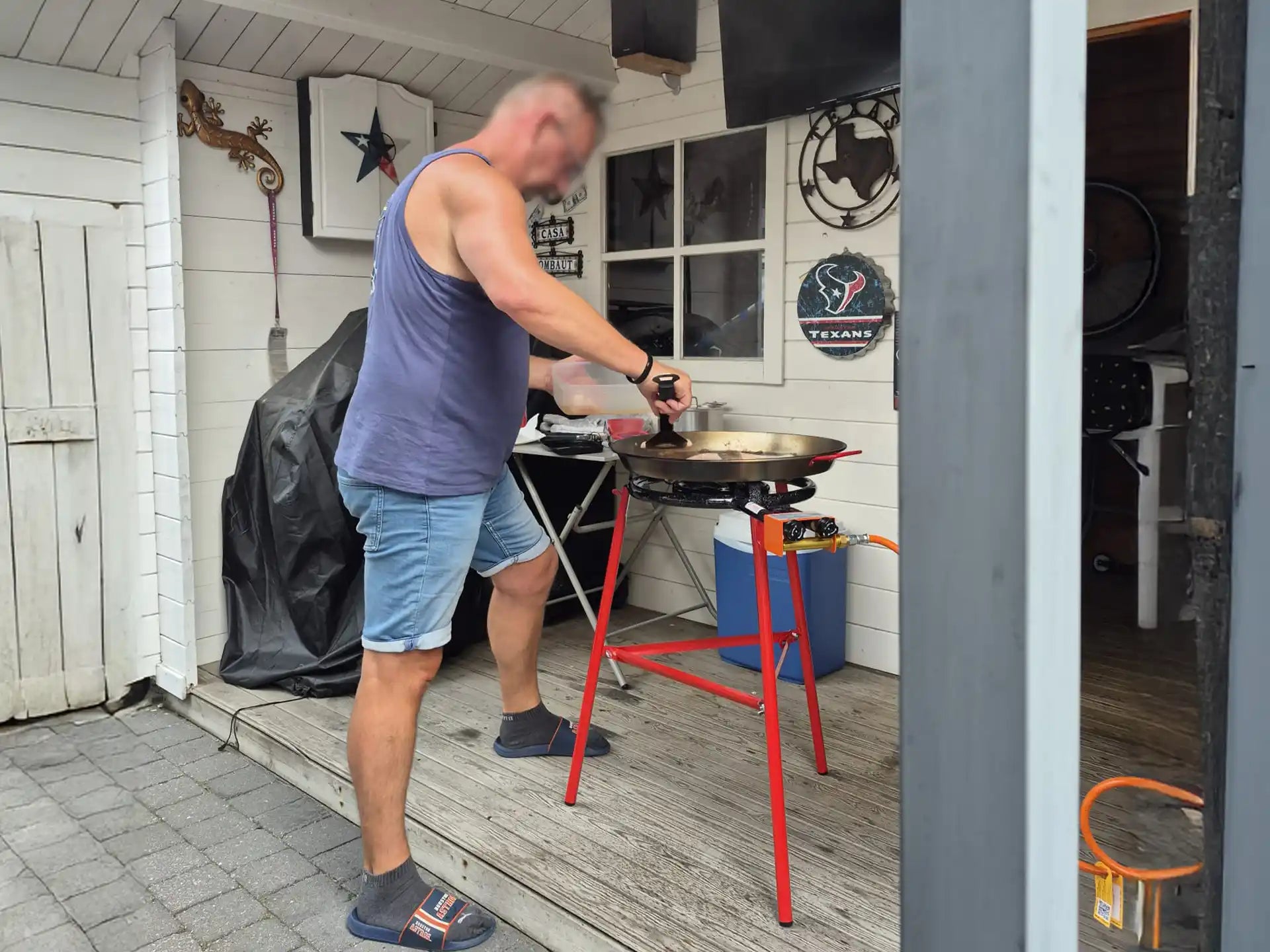 Man in korte broek en hemd bereidt paella in een grote zwarte pan op de rode Garcima paella brander met standaard. De set staat buiten onder een overkapping naast een witte houten wand, met decoratie aan de muur en een gasfles ernaast.