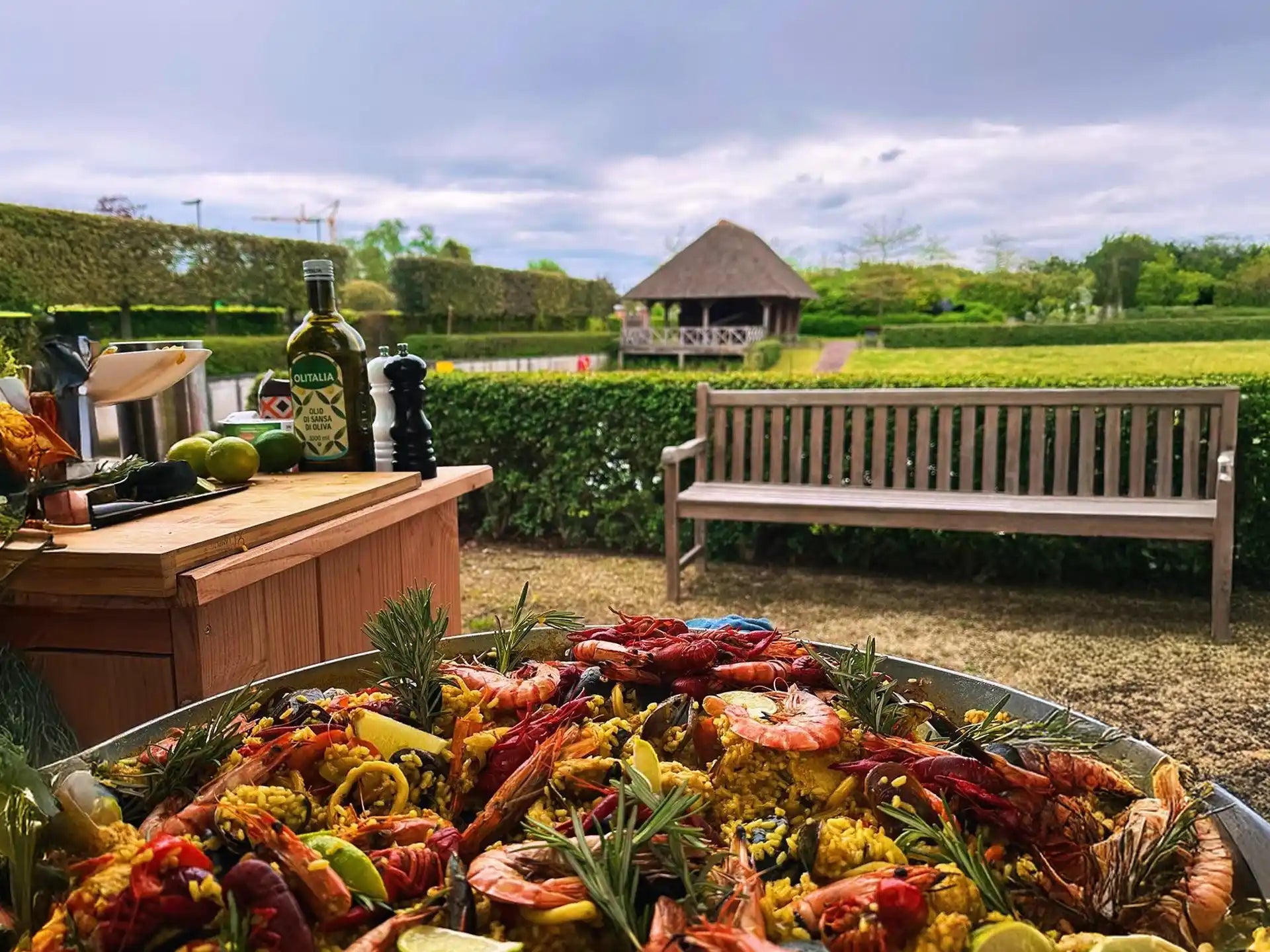Buitenscène met een grote Garcima paella pan gevuld met rijkelijk gegarneerde paella. Op de voorgrond liggen garnalen, mosselen en citroenpartjes. Op de achtergrond staan een houten bank, een tafel met olijfolie en ingrediënten, en een groene tuin met een rieten prieel.