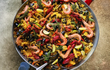 Close-up van een grote Garcima paella pan gevuld met kleurrijke paella. De pan zit vol met garnalen, mosselen, rivierkreeftjes, citroenpartjes en groenten. De pan staat buiten op een stenen ondergrond, naast de voet van een persoon in slippers.