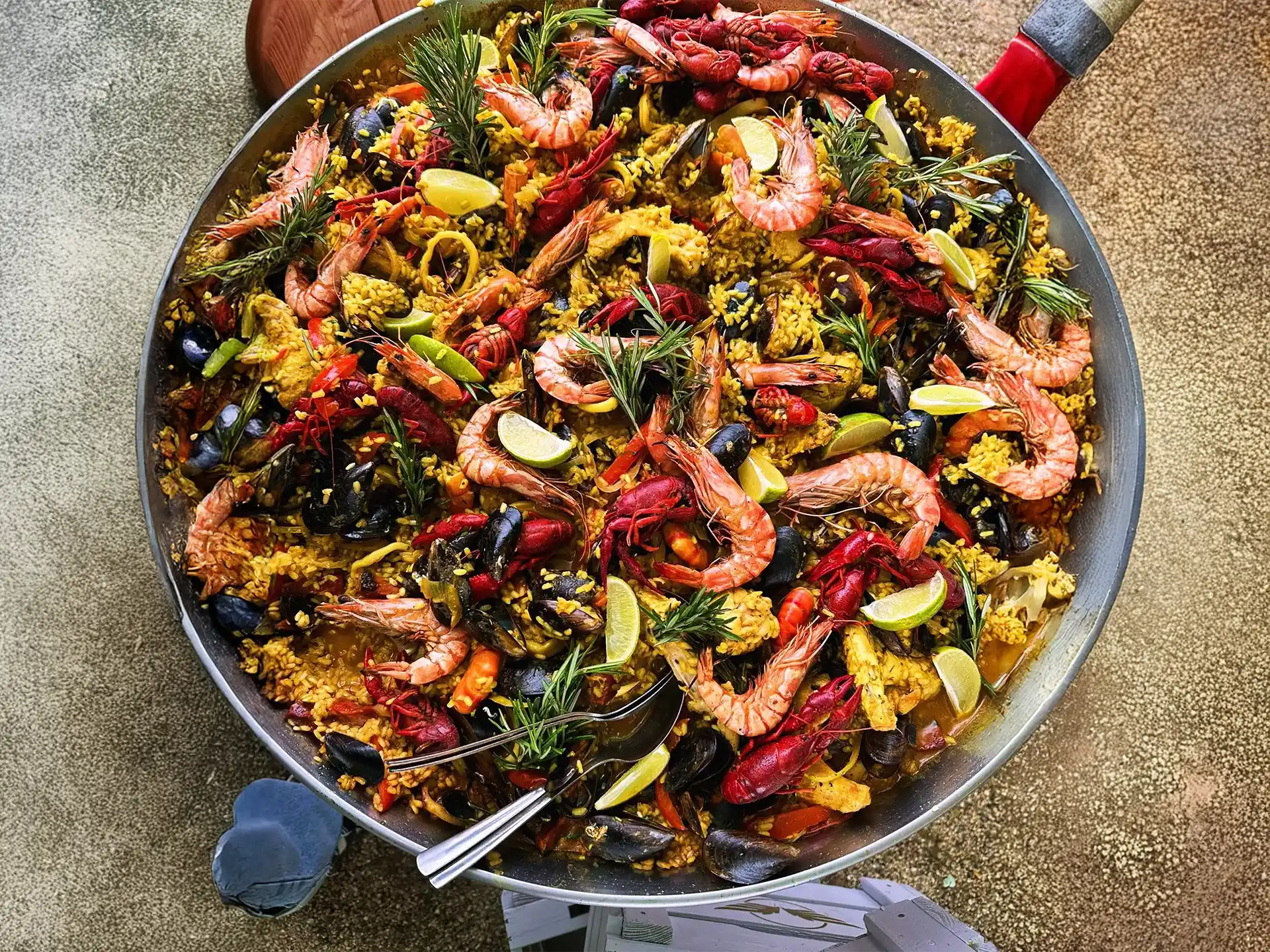 Close-up van een grote Garcima paella pan gevuld met kleurrijke paella. De pan zit vol met garnalen, mosselen, rivierkreeftjes, citroenpartjes en groenten. De pan staat buiten op een stenen ondergrond, naast de voet van een persoon in slippers.