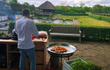 Een kok in witte jas bereidt een gerecht naast een Garcima paella set van 50 cm (Mirador). Op de paella pan worden garnalen gebakken op een brander met rode standaard. De setting is buiten in een verzorgde tuin met vijver, houten bank en rieten prieel op de achtergrond.