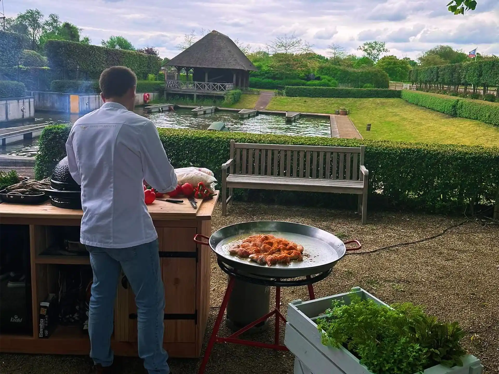 Een kok in witte jas bereidt een gerecht naast een Garcima paella set van 50 cm (Mirador). Op de paella pan worden garnalen gebakken op een brander met rode standaard. De setting is buiten in een verzorgde tuin met vijver, houten bank en rieten prieel op de achtergrond.