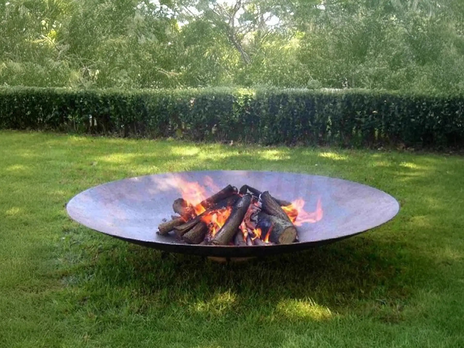 Gofire adeco cortenstaal vuurschaal 150 cm met brandend hout, geplaatst op een grasveld in een ruime tuin met groene heg op de achtergrond.