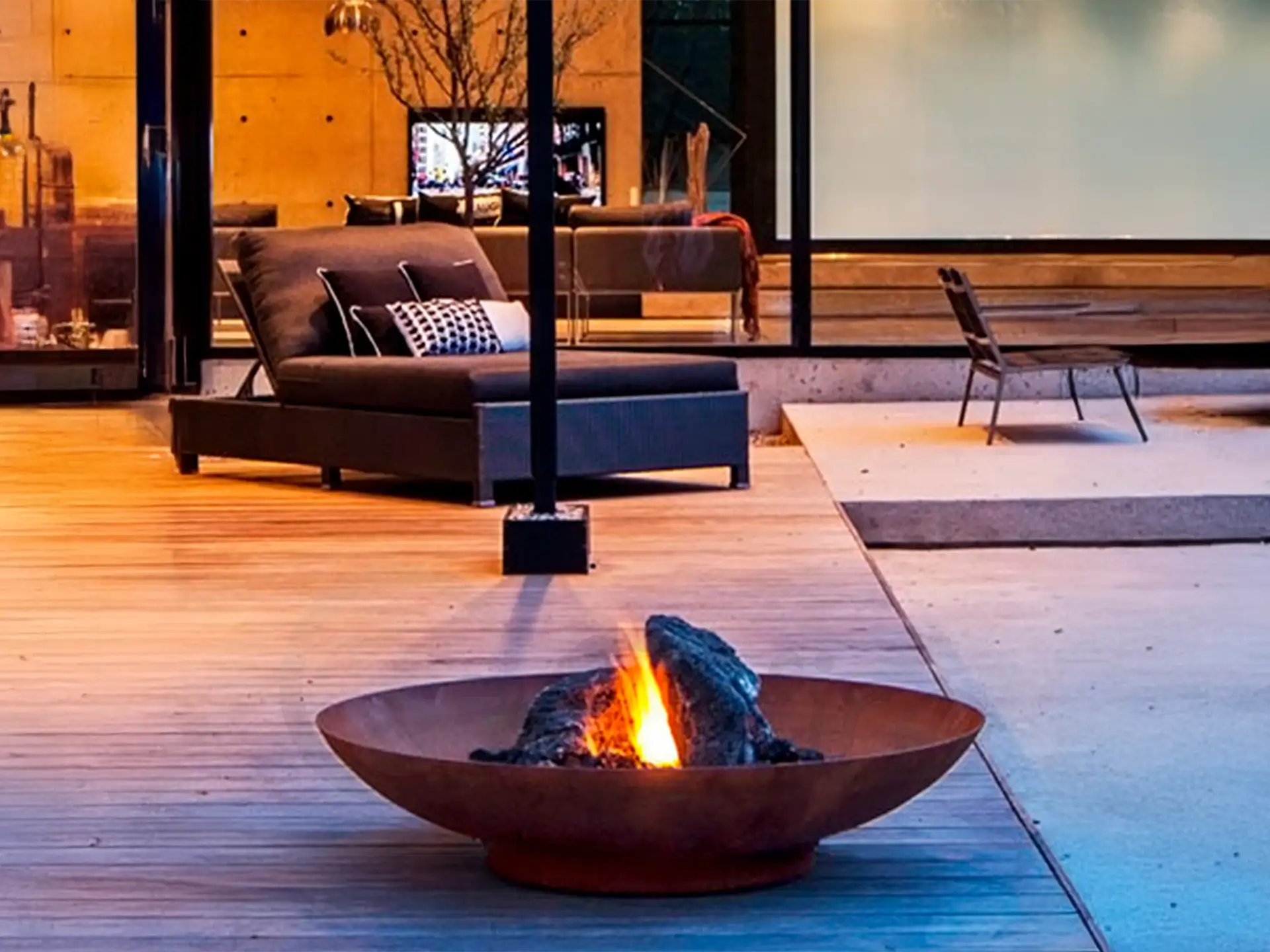 Gofire adeco vuurschaal van cortenstaal met warm vuur, sfeervol geplaatst op een modern houten terras naast een luxe zithoek en grote ramen.