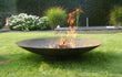 Gofire adeco vuurschaal van cortenstalen met vlammen, brandend op een gazon in een tuin met sierplanten en zwembad op de achtergrond.