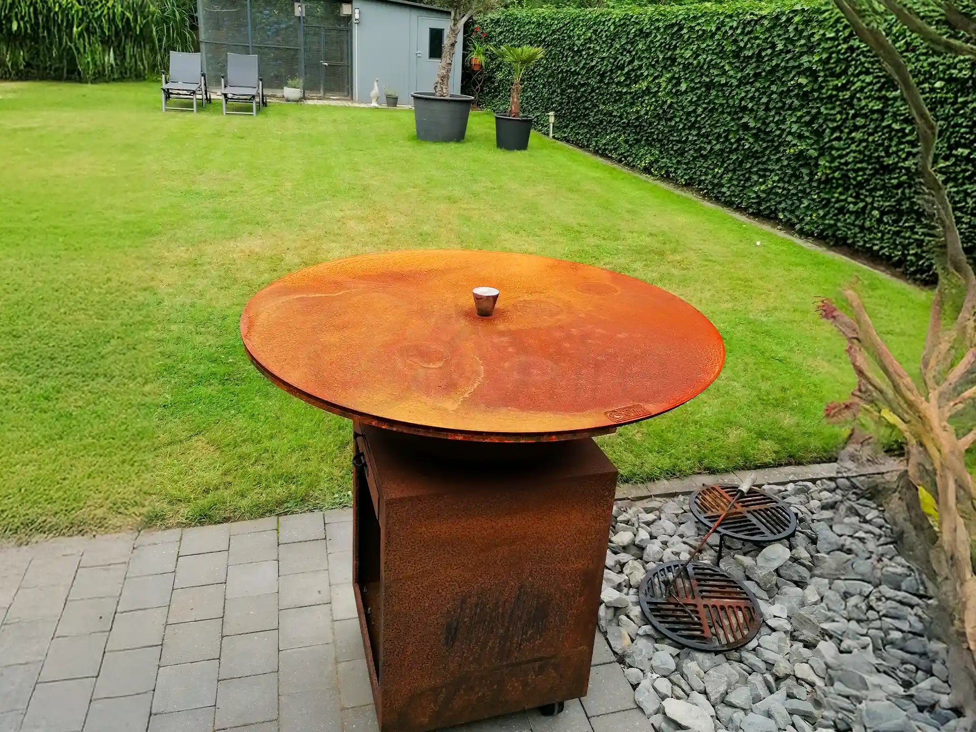 Cortenstalen Gofire deksel voor vuurschaal, geplaatst op een verhoging in een moderne tuin met strak gazon, bestrating en grind, ideaal om de vuurschaal te beschermen en netjes af te dekken.