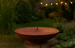 Gofire deksel van cortenstaal voor vuurschaal in een sfeervolle tuin bij schemering, omringd door gras en beplanting, met warme tuinverlichting op de achtergrond die een gezellige ambiance creëert.