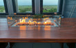 Gofire glasombouw 80 × 30 cm in gebruik op een houten tuintafel met brandende inbouwhaard, moderne setting met uitzicht.