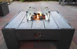 Vierkante Gofire inbouwbrander met glasombouw geïntegreerd in houten tafel, brandend op een terras in daglicht.