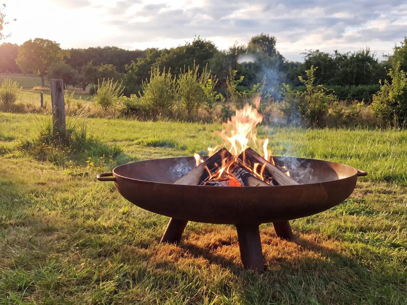 GoFire Lev vuurschaal cortenstaal op poten met brandend houtvuur, in een weide bij zonsondergang.