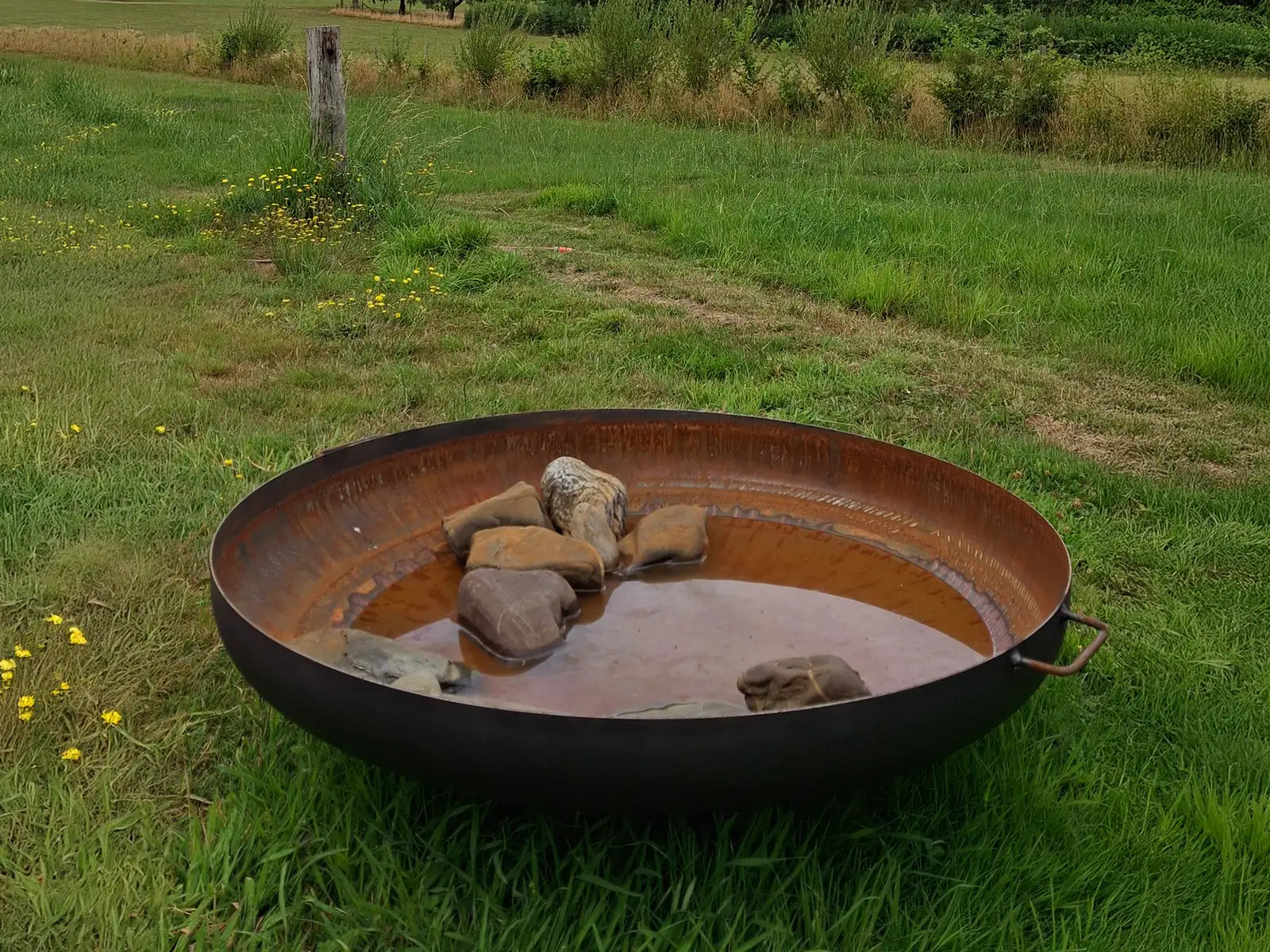 GoFire Lev waterschaal van cortenstaal, deels gevuld met keien als ondiepe drinkplek voor vogels, geplaatst in het gras.