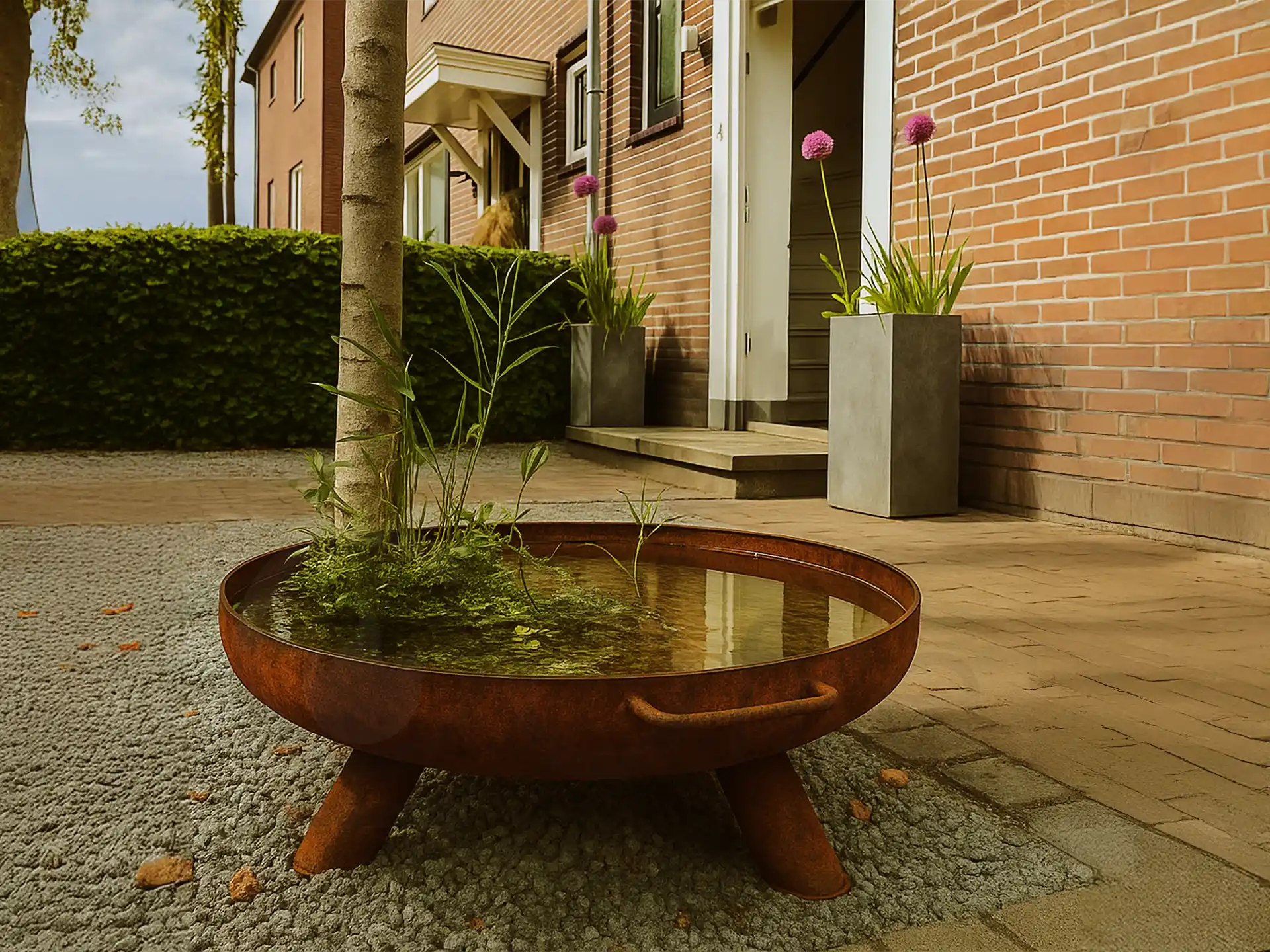 GoFire Lev waterschaal van cortenstaal met waterplanten, geplaatst op een terras naast een boom en grindstrook.