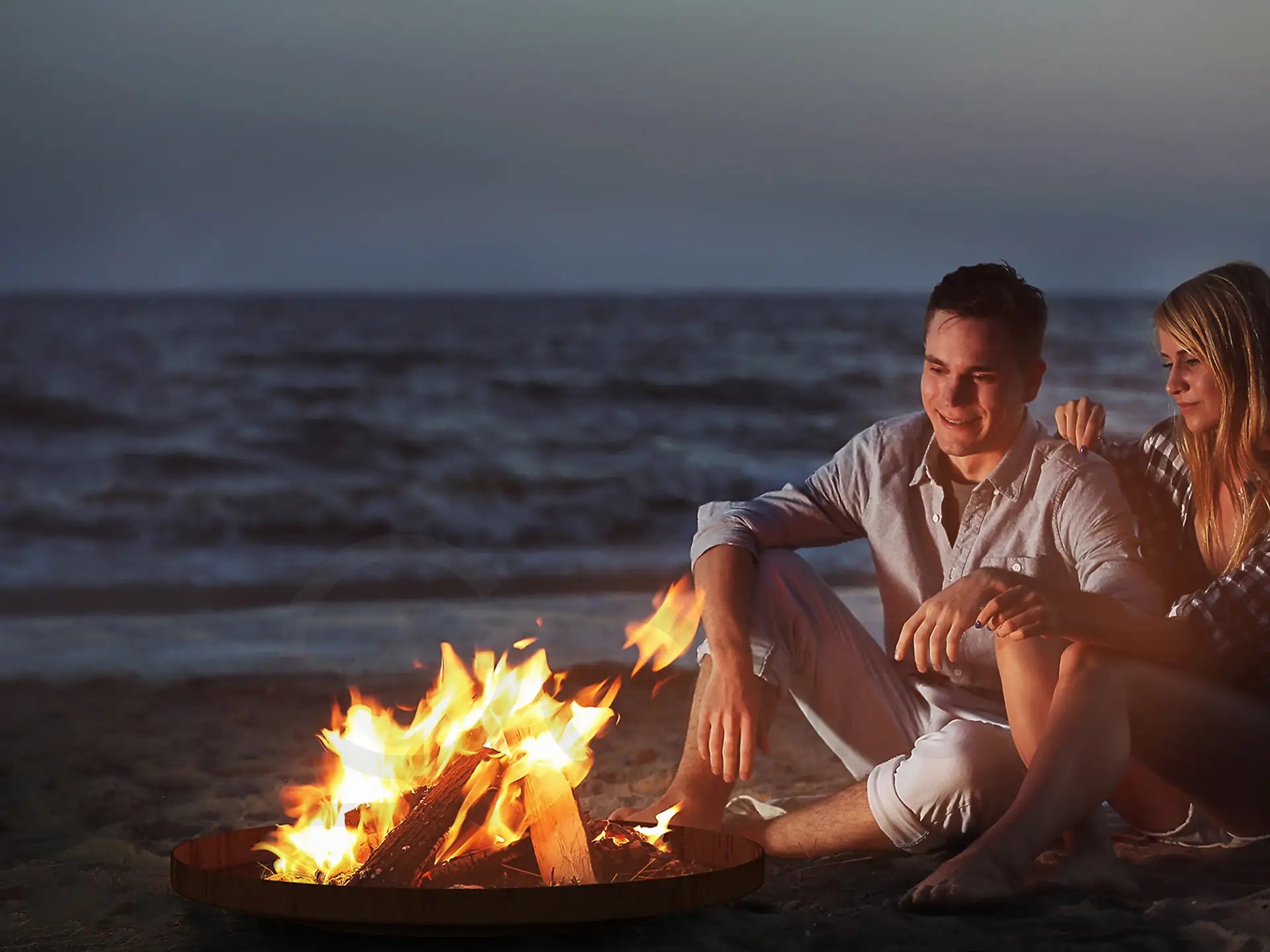 Gofire oda Vuurschaal van cortenstaal met brandend vuur op het strand, waar een stel gezellig bij het warme vuur zit tijdens zonsondergang.