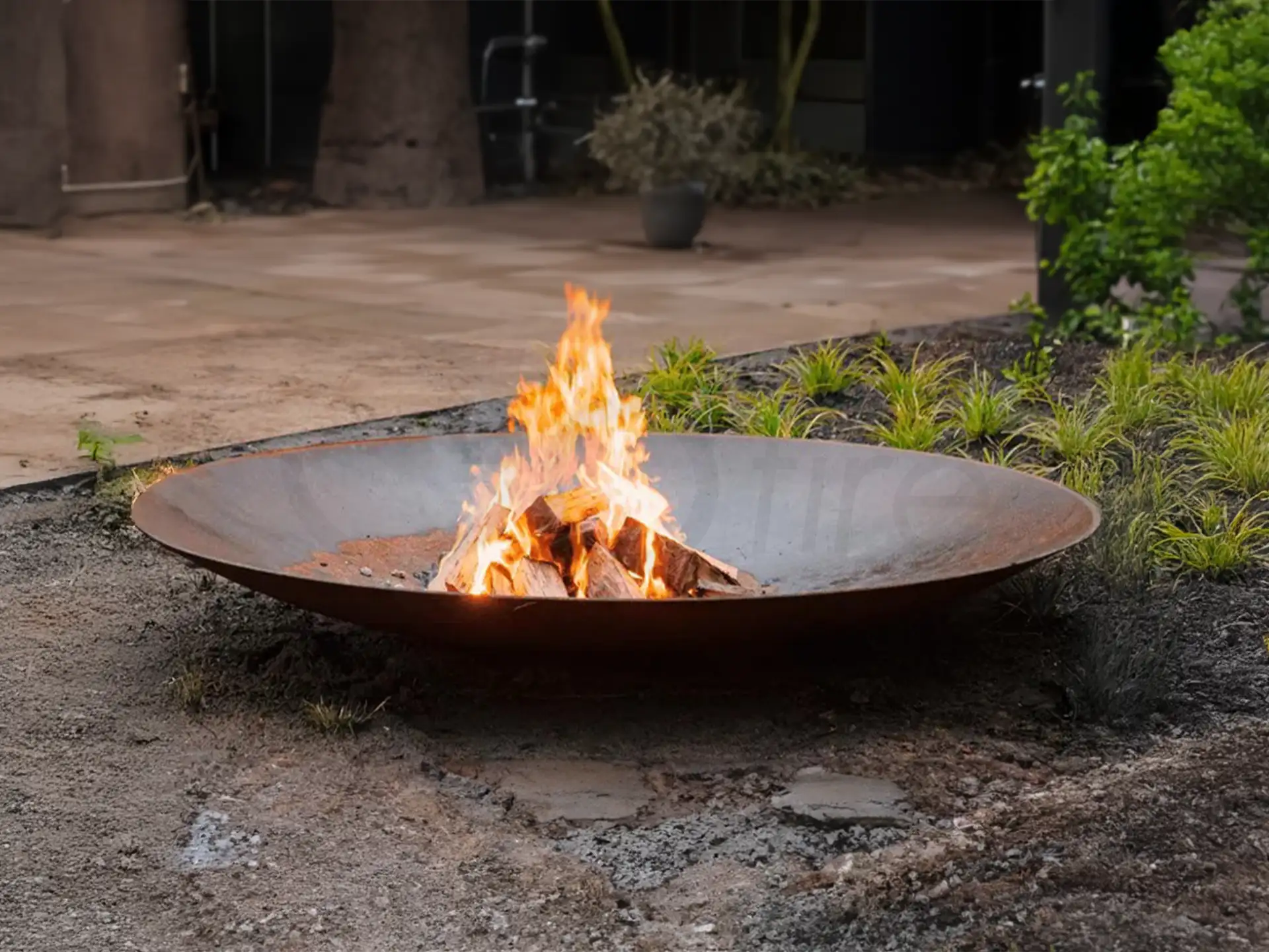 Gofire soho vuurschaal Cortenstalen met knisperend houtvuur, geplaatst in een tuinomgeving met groene planten en moderne bestrating.