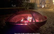 Vuurschaal met fel vuur afgedekt door een zwart vonkenscherm, sfeervol brandend in een tuin met verlichting op de achtergrond.