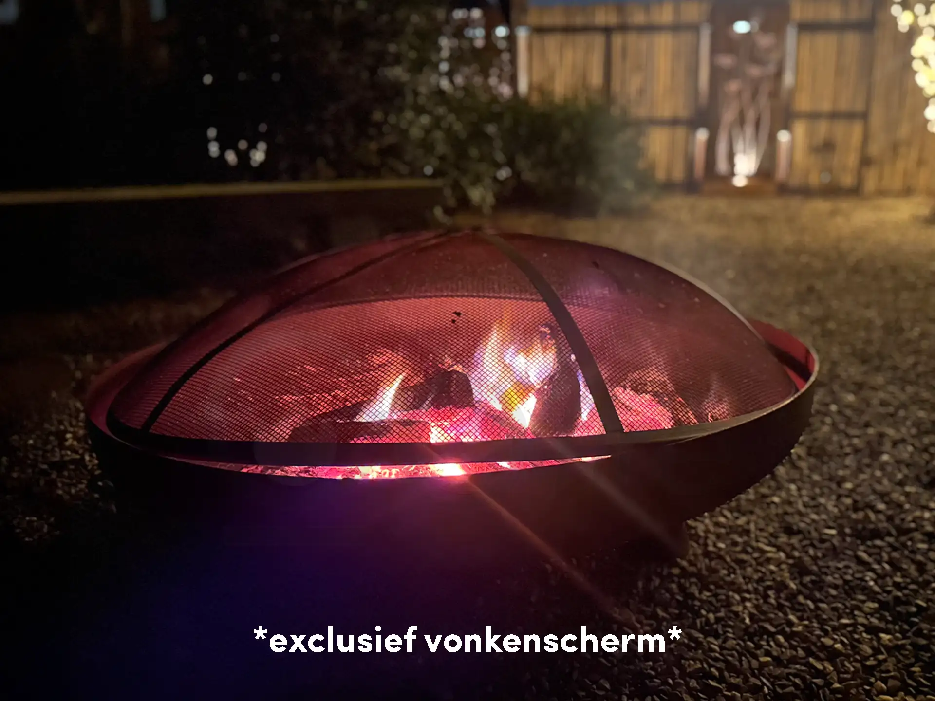 Vuurschaal met fel vuur afgedekt door een zwart vonkenscherm, sfeervol brandend in een tuin met verlichting op de achtergrond.