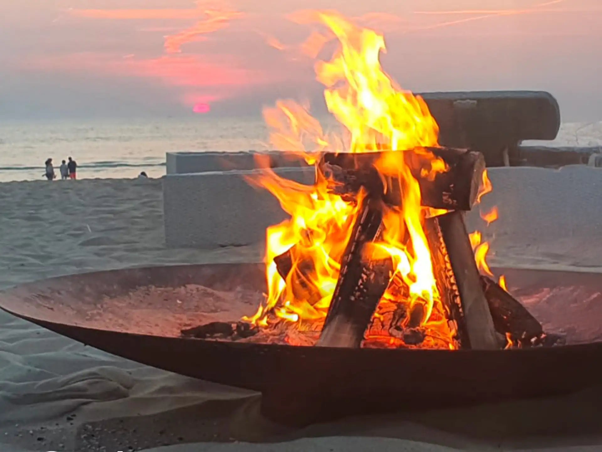 Gofire Soho vuurschaal van cortenstaal met brandend hout op het strand, terwijl de zon ondergaat en er een warme sfeer ontstaat bij de zee.