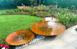Set van GoFire Soho waterschalen van cortenstaal met fonteintjes, geplaatst naast elkaar in een moderne tuin met gazon en sierplanten.
