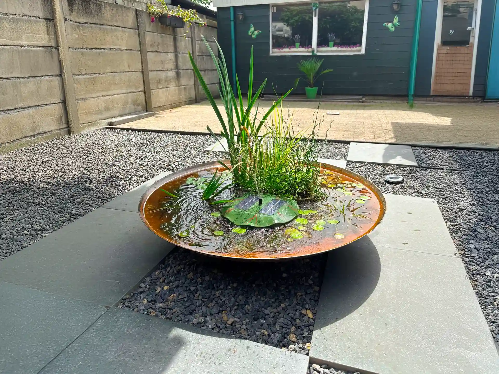 Gofire waterschaal gevuld met waterplanten en fontein, geplaatst op een modern terras met grind en tegels voor een decoratief waterelement.