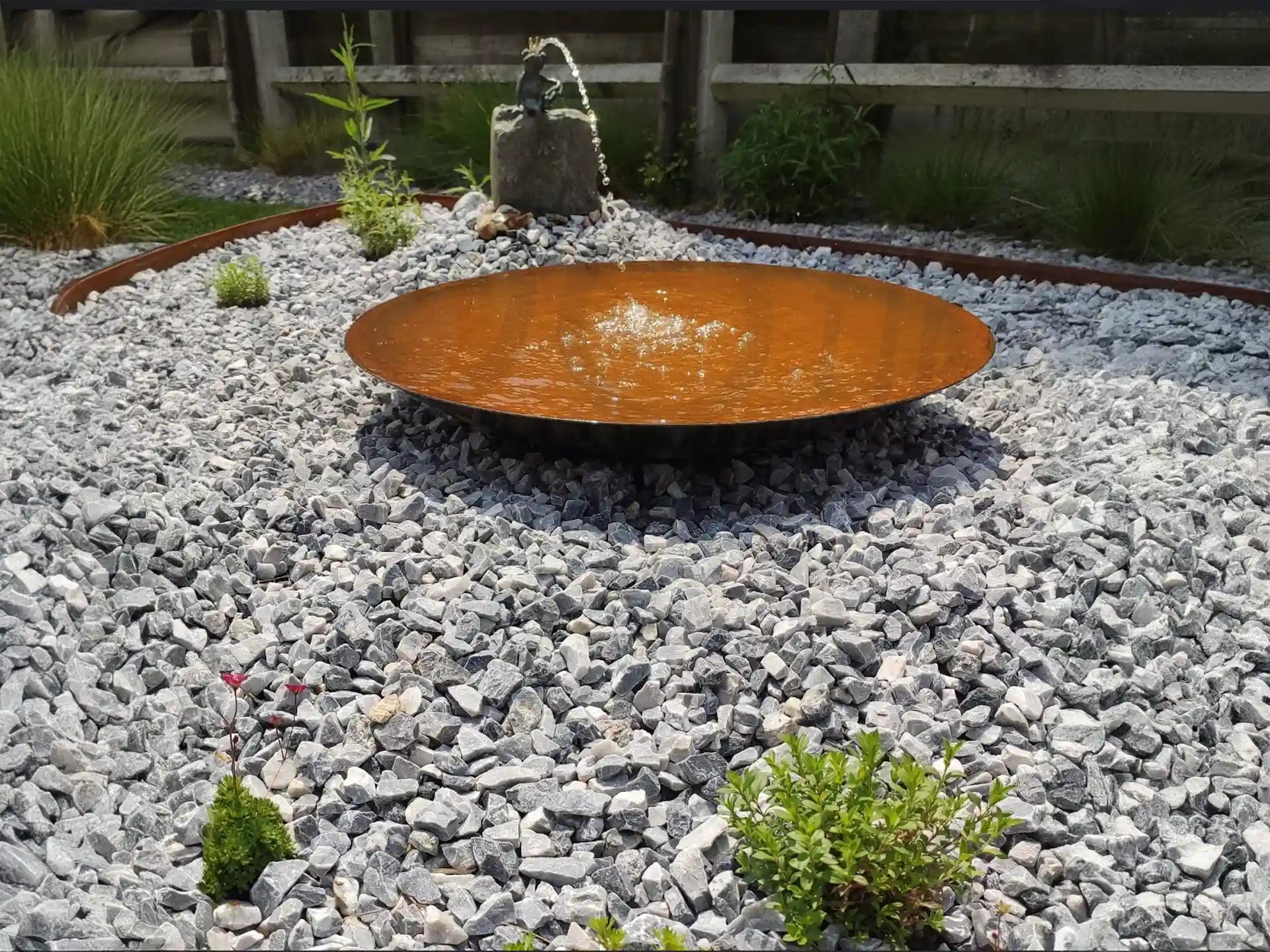 GoFire Soho waterschaal van cortenstaal met fontein, stijlvol geplaatst in een grindtuin met groene accenten en siergras.