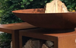 Close-up van de Halo cortenstalen vuurschaal gevuld met houtblokken, rustend op een robuuste houtopslag van cortenstaal in een groene tuinsetting.
