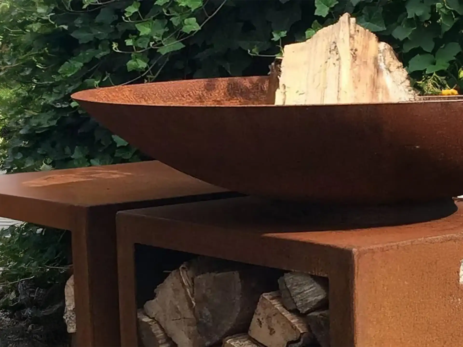 Close-up van de Halo cortenstalen vuurschaal gevuld met houtblokken, rustend op een robuuste houtopslag van cortenstaal in een groene tuinsetting.
