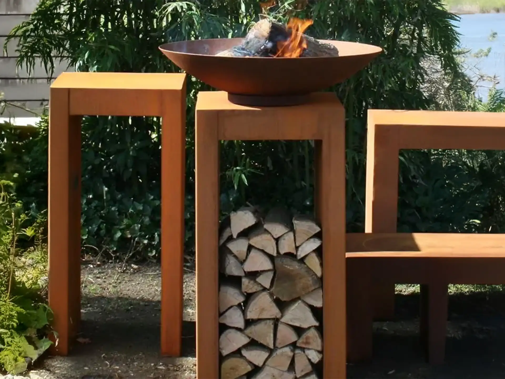 Close-up van de Halo cortenstalen vuurschaal gevuld met houtblokken, rustend op een robuuste houtopslag van cortenstaal in een groene tuinsetting.