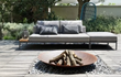 Halo Cortenstalen vuurschaal met houtblokken, geplaatst op een terras met een moderne loungebank en sfeervolle tuinbeplanting eromheen.
