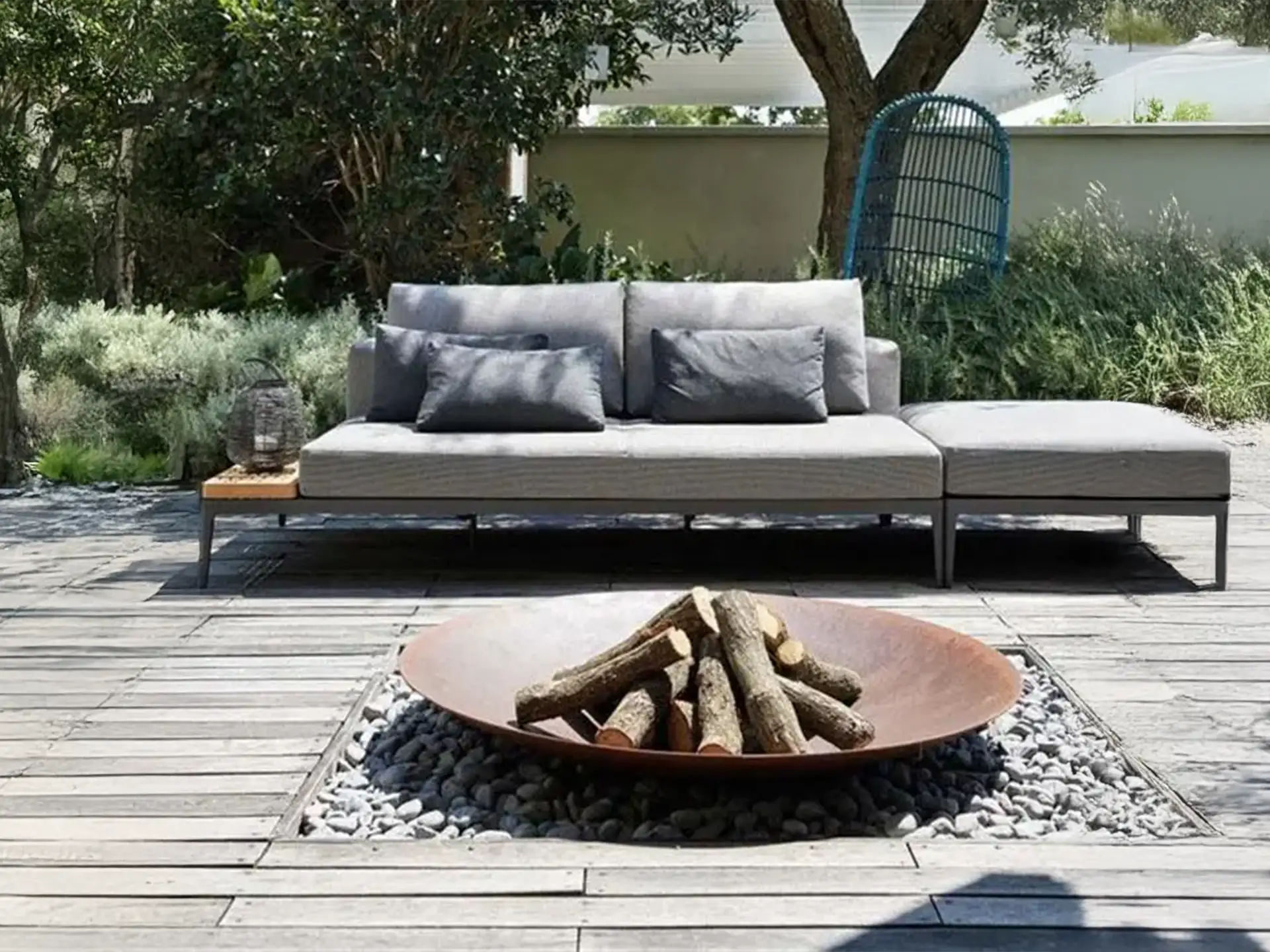 Halo Cortenstalen vuurschaal met houtblokken, geplaatst op een terras met een moderne loungebank en sfeervolle tuinbeplanting eromheen.