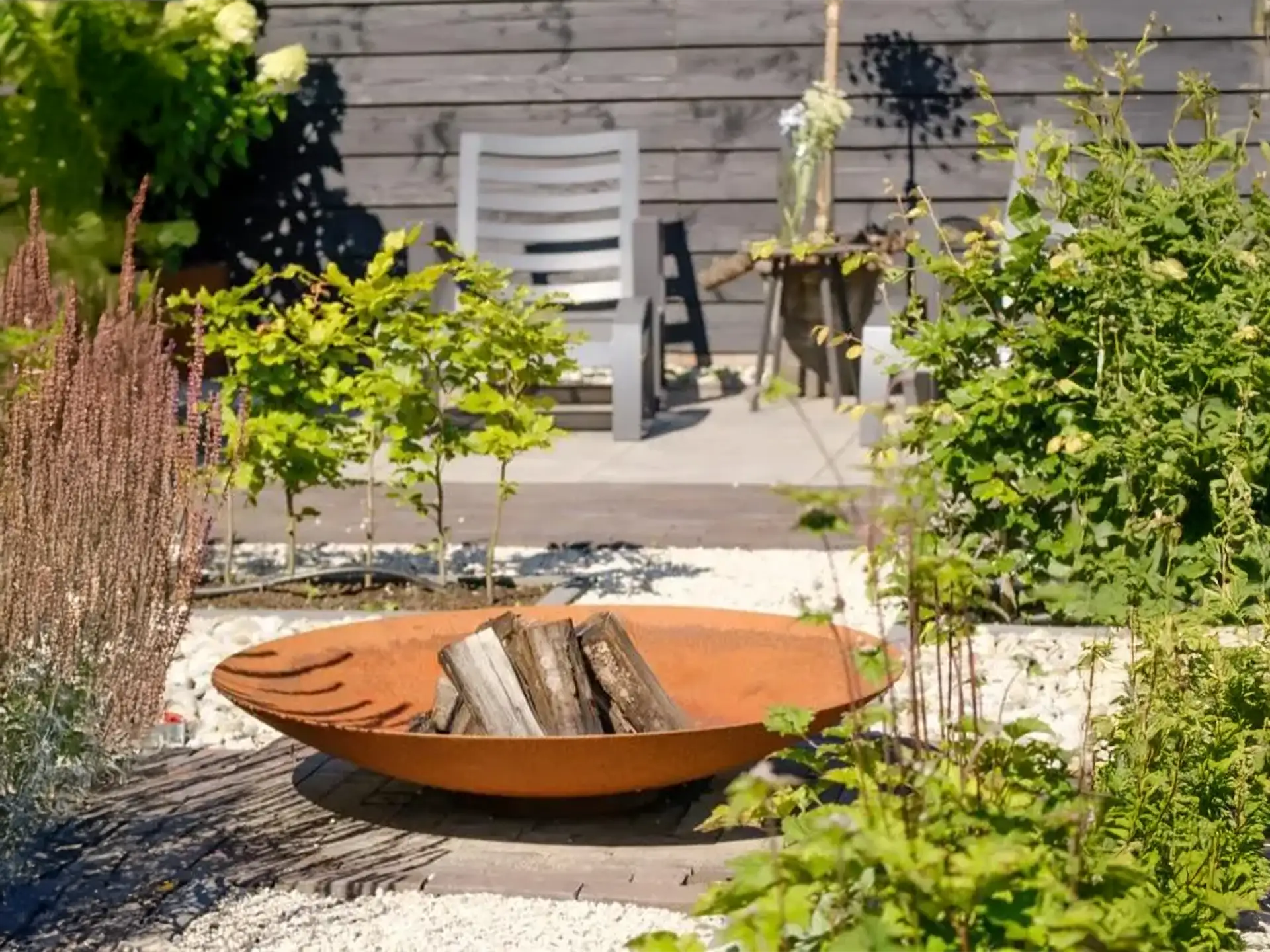 Halo Cortenstalen vuurschaal geplaatst in een moderne tuin, gevuld met houtblokken en omringd door groene planten en siergrassen.