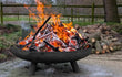 Lev vuurschaal zwart met brandend houtvuur en gloeiende houtblokken, geplaatst op poten in een natuurlijke buitenomgeving met houtopslag op de achtergrond.