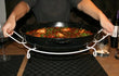 Onderzetter voor paella pan Deluxe in gebruik, persoon tilt pan vol paella van de tafel met stevige standaard.