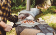 Ooni Karu 12 pizza oven in gebruik buiten, persoon schuift pizza in de hout- of houtskoolgestookte oven met pizzaschep, sfeervolle setting.