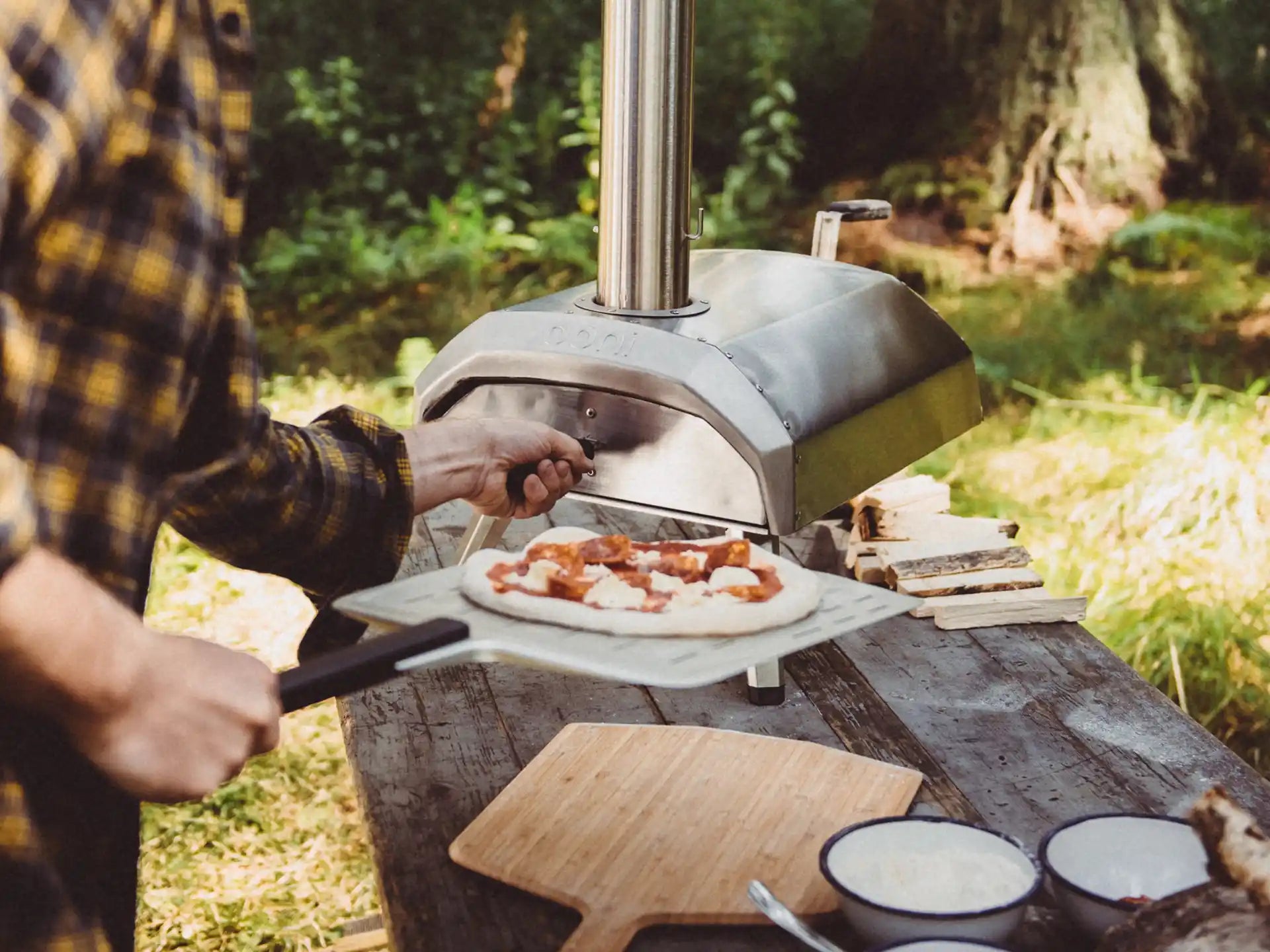 Ooni Karu 12 pizza oven in gebruik buiten, persoon schuift pizza in de hout- of houtskoolgestookte oven met pizzaschep, sfeervolle setting.