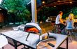 Ooni Karu 12 pizza oven hout of houtskoolgestookt, in gebruik buiten op tafel met pizza in de oven en mensen op de achtergrond.