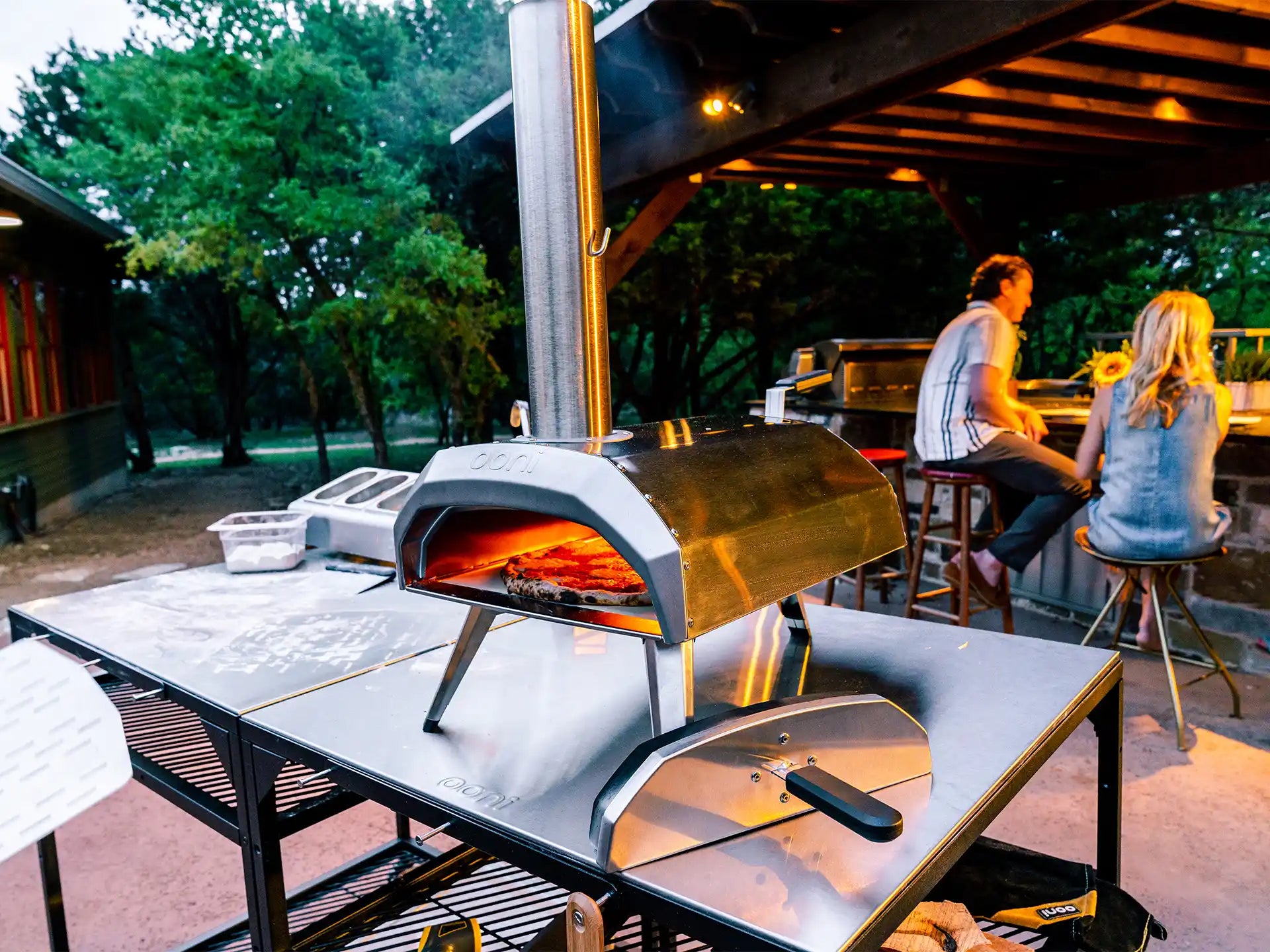Ooni Karu 12 pizza oven hout of houtskoolgestookt, in gebruik buiten op tafel met pizza in de oven en mensen op de achtergrond.