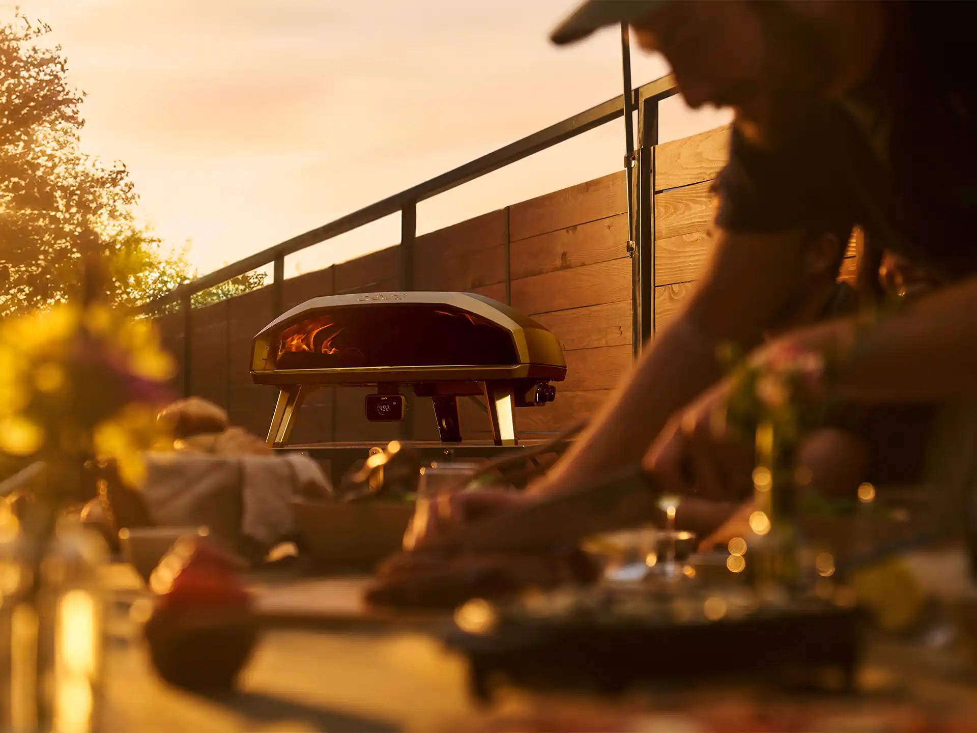 Ooni Koda 2 Max pizza oven buiten in gebruik tijdens een sfeervol etentje, persoon bereidt pizza naast de oven.