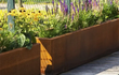 Grote rechthoekige Adezz Carrez Cortenstaal Plantenbak met wielen, beplant met bloemen en groen langs een moderne tuin.